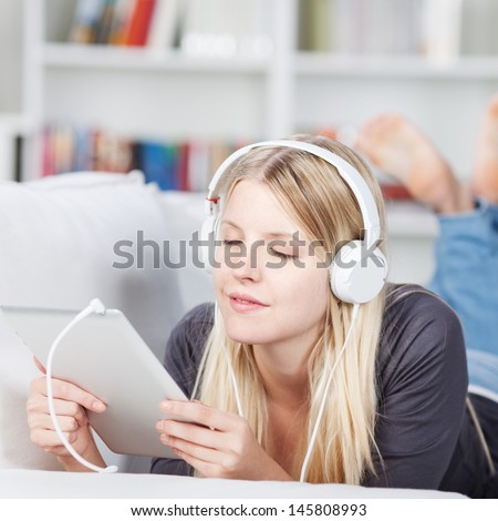Young woman enjoying music on headphones using digital tablet while lying on sofa at home