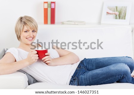 Pretty young blond woman relaxing on a sofa with a red mug of coffee or tea in her hands smiling at the camera