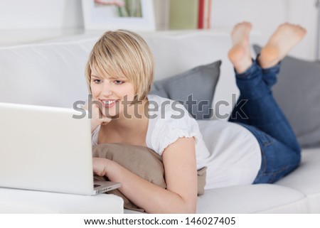 Woman relaxing on a sofa with a laptop computer balanced on the arm reading the information on the screen with a pleased smile