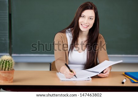 Beautiful young trainee female teacher at her desk in front of the blackboard writing notes and smiling at the camera