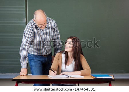 Pretty young woman university student with a middle-aged male lecturer or tutor leaning over her at her desk to help her with her project against a green blackboard