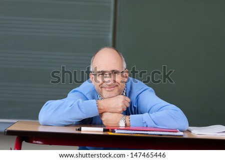 Friendly male teacher relaxing in the classroom in front of the blackboard with his chin resting on his hands on the desk