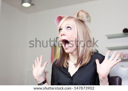 Shocked woman wearing large hair curlers in her hair at a hairdresser raising her hands in mock horror in a fun portrait