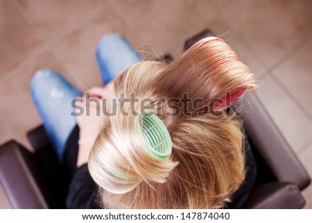 Overhead view of a woman with large curlers in her blond hair to give it lift sitting in an armchair in a hair salon