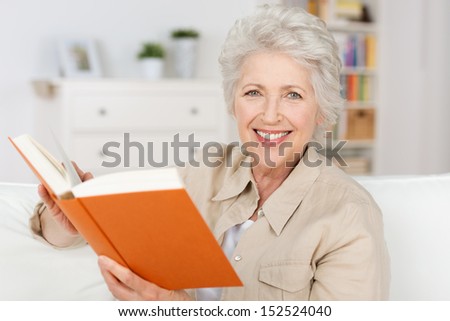 Smiling elderly lady reading a book while sitting in her living room pausing to look and smile at the camera