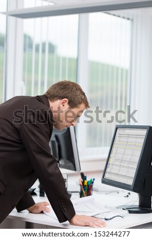 Businessman reading information on the computer as he stands leaning over his desk at the office in front of a large window