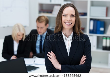 Confident attractive young businesswoman standing with folded arms in a busy office smiling at the camera