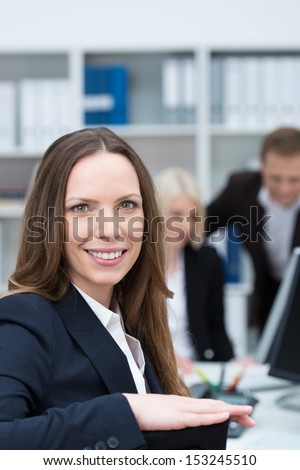 Smiling attractive young businesswoman in a busy office turning in her chair to look at the camera