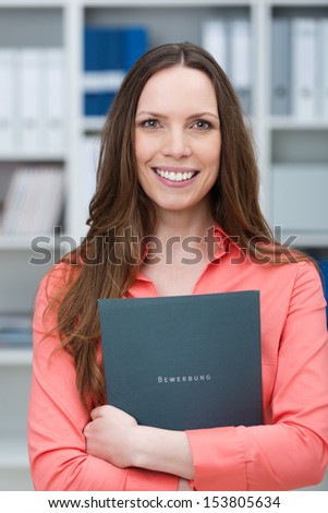 Smiling young job applicant clutching a folder with her curriculum vitae to her chest standing in an office