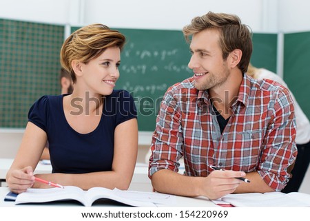 Attractive young male and female college students sharing a desk together sit in the classroom discussing a project and smiling at each other
