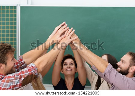 Successful teamwork in the classroom with a group of diverse college or university students standing in a circle all raising their hands to meet in the centre