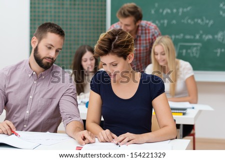 Two young university students, a bearded man and attractive young woman, sitting at their desks sharing information and helping each other