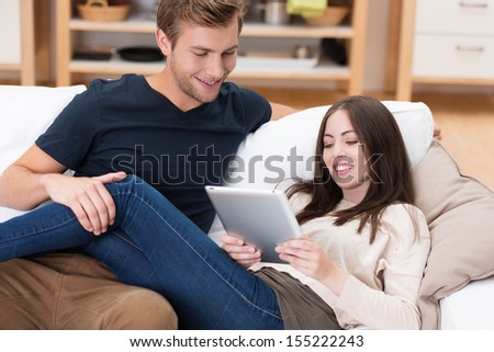 Young man and woman relaxing at home lying on the sofa together smiling and reading information on a tablet computer