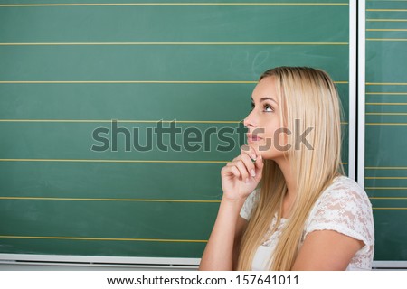 Beautiful thoughtful young student or teacher standing in front of a black green chalkboard with her hand to her chin staring pensively into the air
