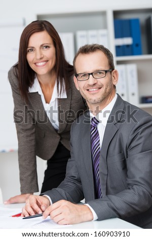 Smiling confident business manager with his pretty young female secretary leaning over his shoulder as he sits at his desk in the office