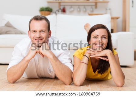 Lazy attractive young couple unwinding at home lying on their stomachs on the wooden floor in the living room grinning at the camera