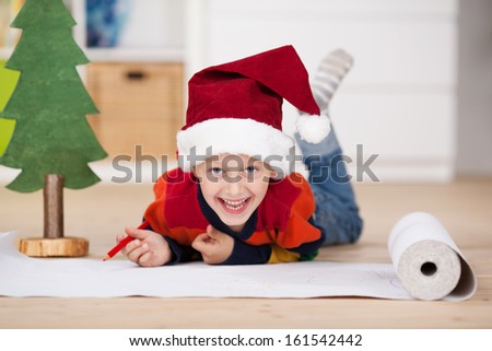 Playful laughing young boy in a Santa Hat lying on the floor drawing on a length of paper alongside a Christmas tree decoration