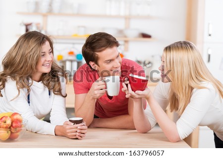 Three happy young college students having coffee together in the kitchen laugh and smile as they group around the wooden counter