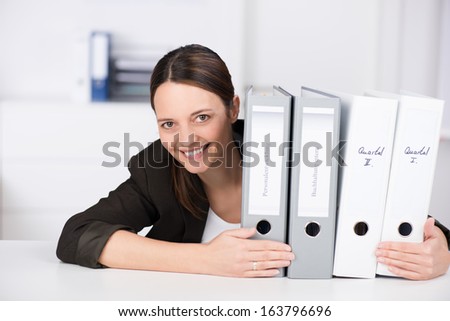 Happy attractive businesswoman sitting at a table with a row of office files peering around the side with a cheerful friendly smile