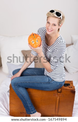 Elated trendy l young blond woman with her piggy bank sitting on her suitcase grinning at the camera with satisfaction as she plans to fund her travels from her savings