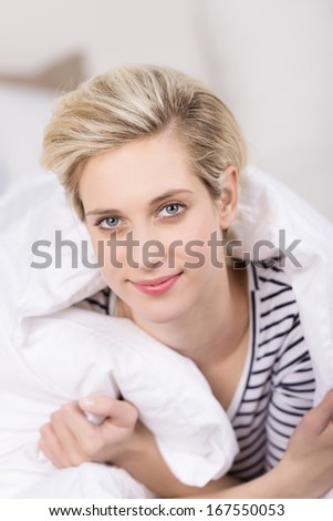 Beautiful young woman with a stylish blond hairstyle having a lazy day smiling as she cuddles up in a warm duvet