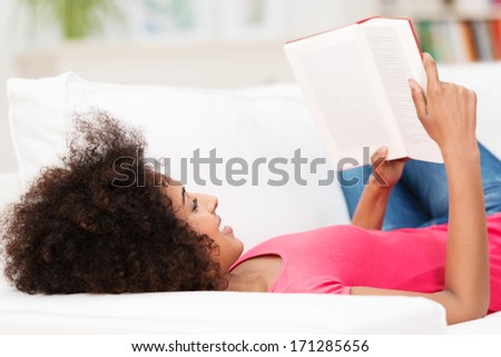 Young African American woman smiling while laying comfortably on her back on the sofa, reading a book