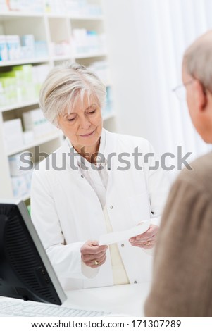 Female pharmacist checking a prescription behind the counter watched by a patient