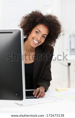 Cute African American businesswoman with a wild afro hairdo and friendly smile peering around her computer monitor at the camera