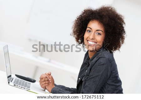 Happy contented young African American woman with an afro hairstyle sitting at her desk in front of a laptop computer turning to smile at the camera