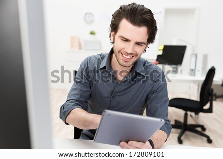 Smiling businessman surfing the internet in the office on a handheld tablet computer using his finger to activate the touchscreen