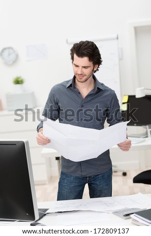 Businessman standing behind his desk in the office looking at a report or architectural or engineering drawing which he is holding open in his hand