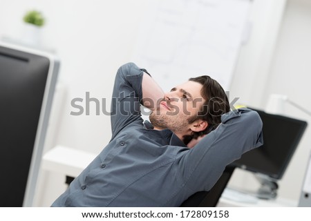 Businessman sitting daydreaming at his desk at the office leaning back in his chair looking up into the air with a pleased thoughtful smile and his hands clasped behind his head