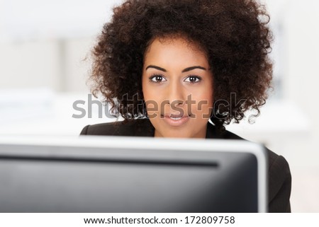 Beautiful serene African American businesswoman with a wild afro hairstyle sitting looking over the top of her monitor at the camera with a quiet smile