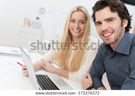 Young couple or business partners working together at a laptop in an office turning to give the camera warm friendly smiles