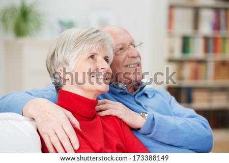 Happy senior couple sitting together arm in arm on a couch in the living room looking upwards watching something