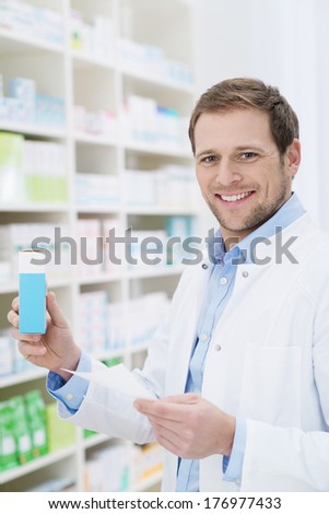 Smiling pharmacist holding a blank box of medication as he stands at the shelves in the pharmacy fulfilling a prescription
