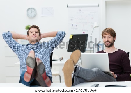 Two relaxed confident young business partners sure of their own success sitting with their feet up on the desk and complacent smiles