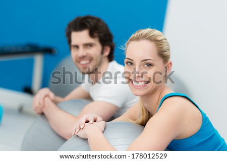 Smiling attractive couple doing pilates exercises in a gym together working out with large gym balls and pausing to smile at the camera with focus to the womans face