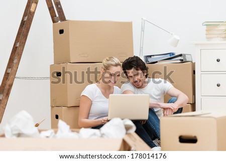 Young couple surrounded by packing boxes smiling as they use their laptop to make contact with friends while moving house