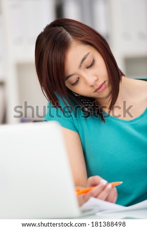 Beautiful young Asian woman concentrating on her work sitting at her desk behind her laptop reading some paperwork with a pen in her hand