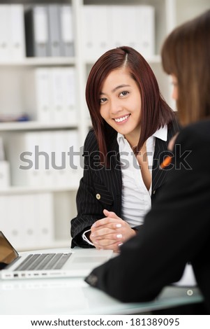 Two businesswomen working together as a team in the office sitting at a desk sharing a laptop with focus to a friendly Asian woman facing the camera with a smile