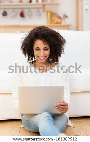 Smiling young African American woman with a wild curly afro hairstyle at home sitting in casual jeans on the floor in the living room using a laptop computer