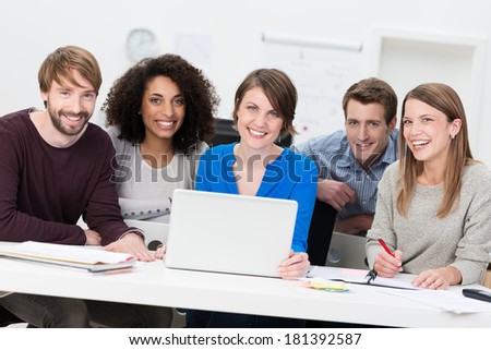 Happy successful young multiethnic business team sitting together in a meeting at a table in the office working on a laptop computer smiling at the camera