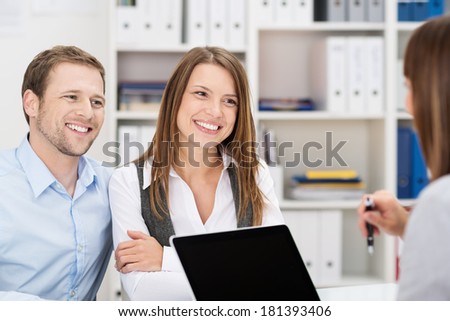 Smiling attractive young couple sitting in an office chatting to a business broker, insurance agent or adviser
