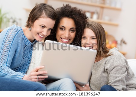 Three beautiful women sitting close together on a sofa in the living room smiling as they read the screen on a laptop computer