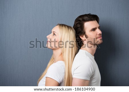 Attractive young couple standing back to back both staring thoughtfully into the distance with quiet smiles, on grey