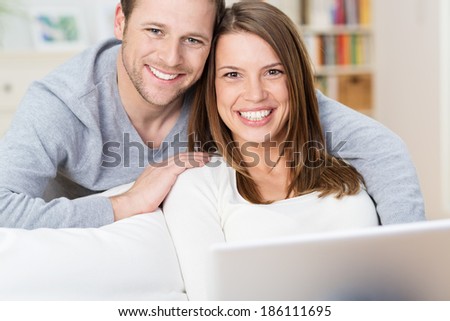 Smiling young couple sharing a laptop computer as they sit on a couch at home surfing the internet together and looking up to smile at the camera