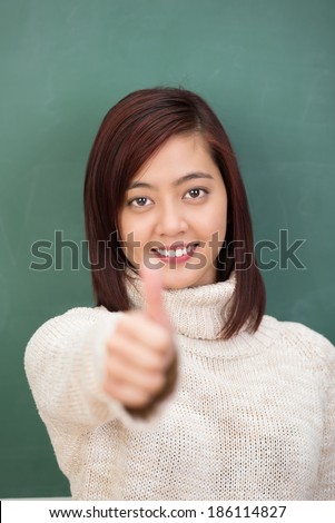 Pretty young asian student in a stylish polo-neck sweater giving a thumbs up gesture of approval as she gives a contented smile of satisfaction