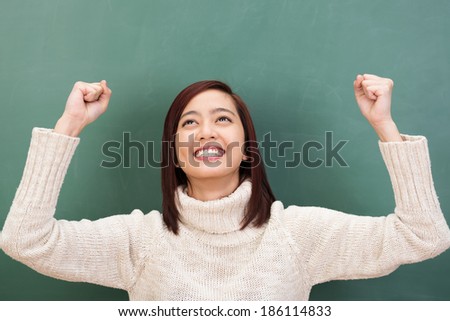 Jubilant attractive young Asian student cheering in elation raising her fists in the air in her excitement against a green chalkboard