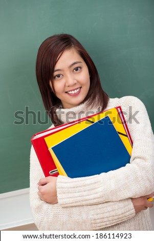 Pretty young Asian student clasping a batch of brightly coloured files in her arms as she stands in front of the blackboard